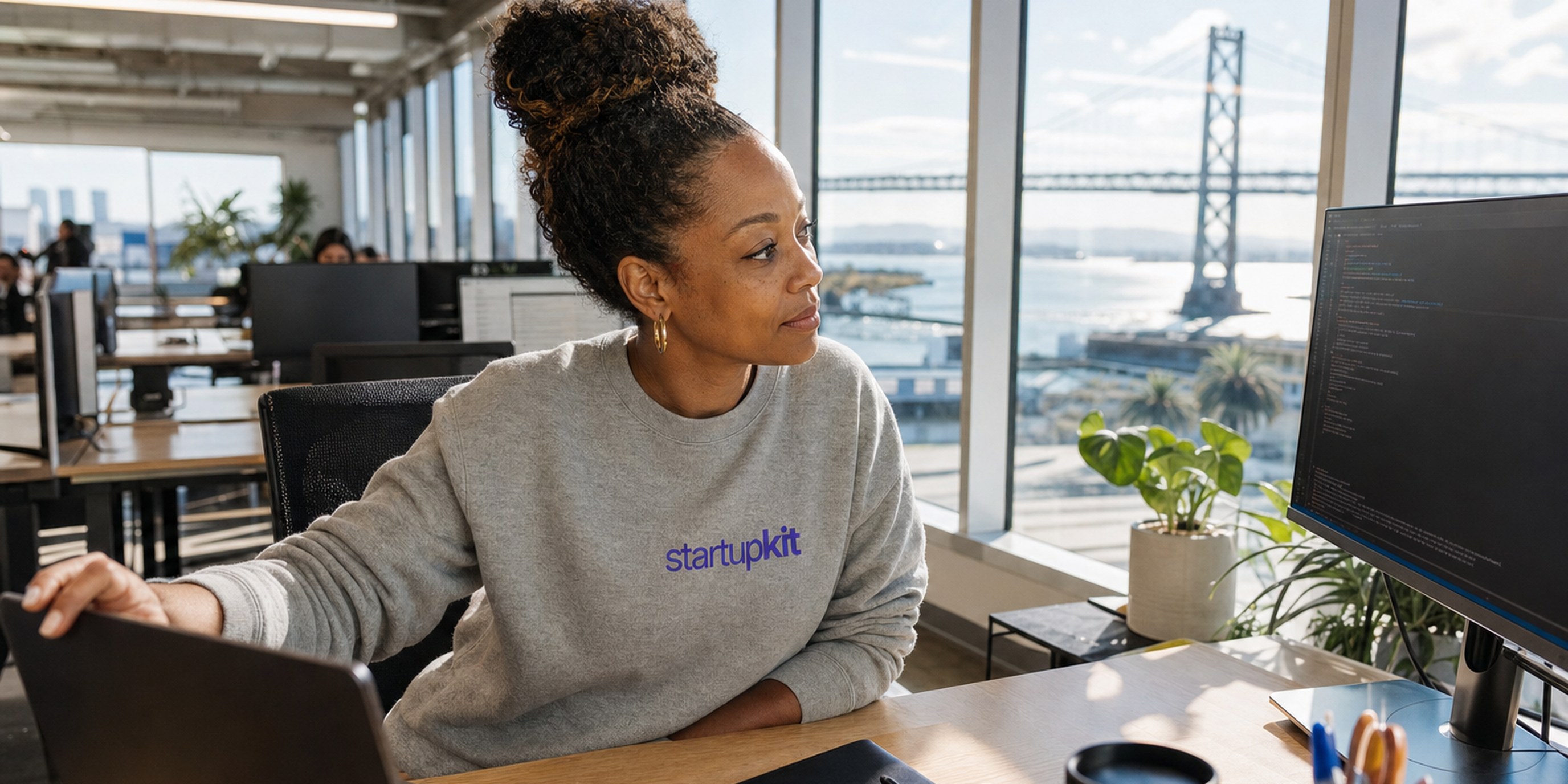 A recruiter at a co-working desk closing her laptop on an overflowing application inbox, turning instead to review a candidate's completed work sample on a second monitor