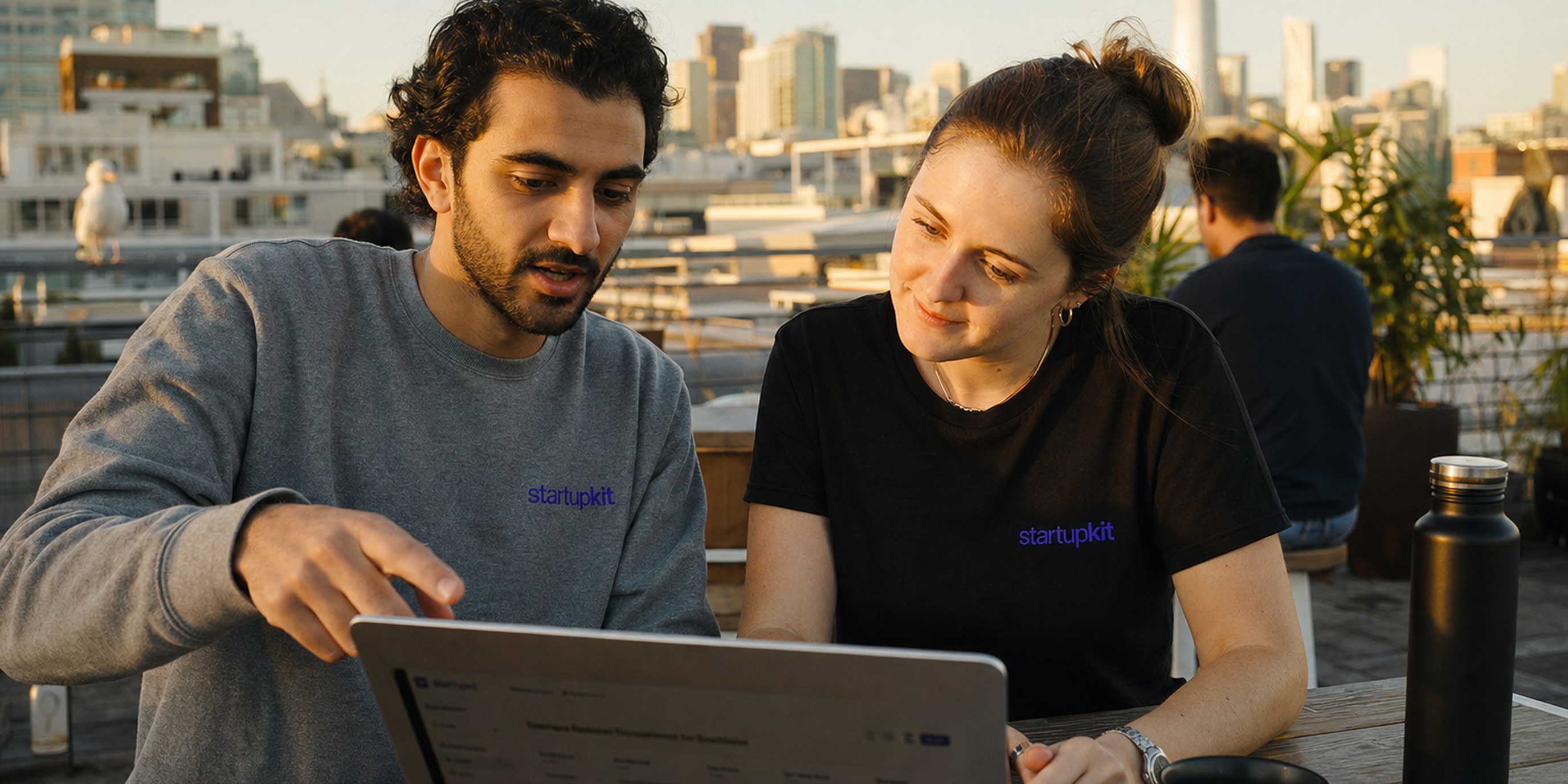A young hiring duo, a Middle Eastern man and a white woman in their late twenties, collaborating over a laptop showing a hiring pipeline on a sunny San Francisco rooftop co-working deck at golden hour, the city skyline behind them