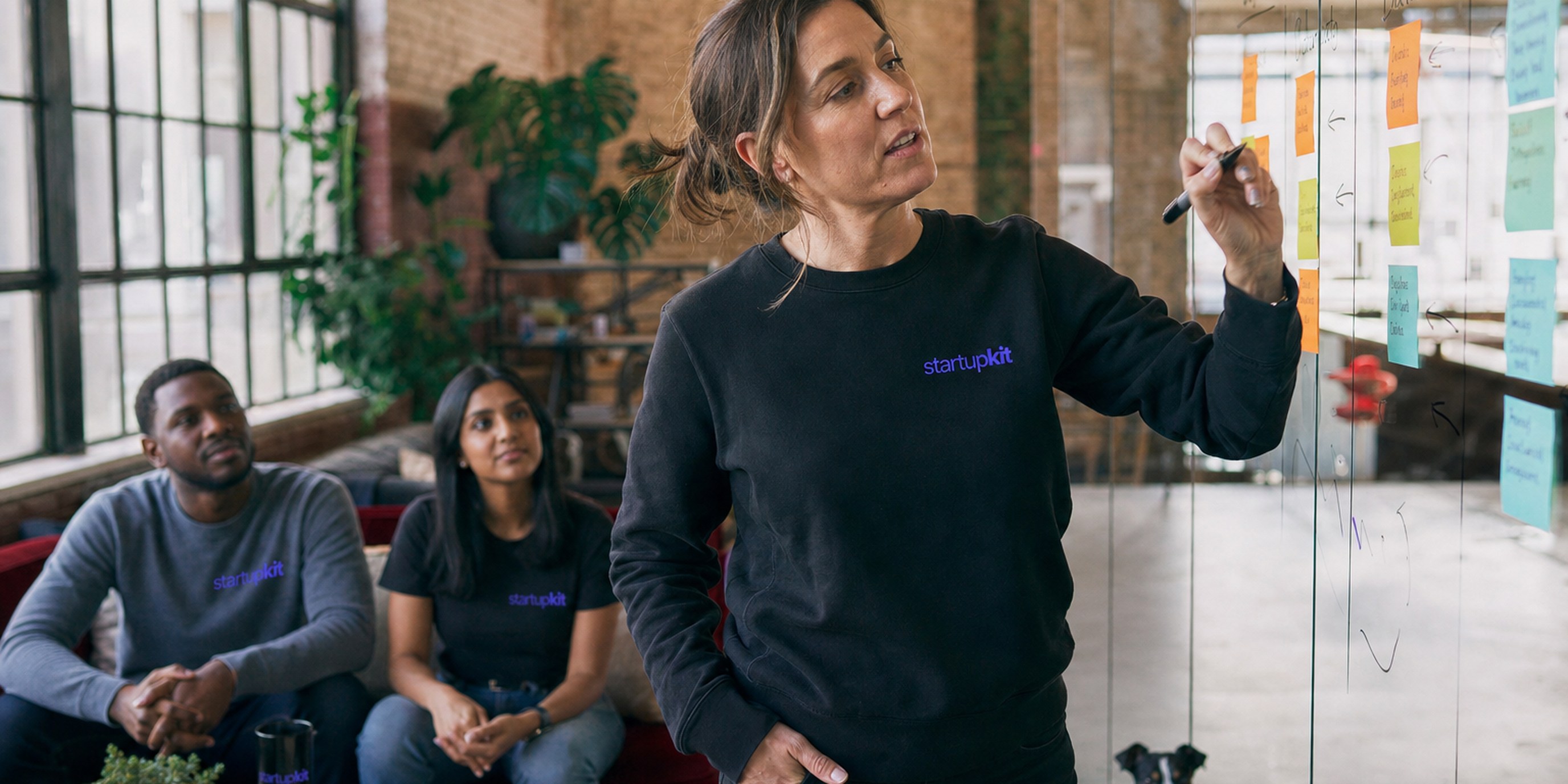 A small startup trio led by a woman gathered around a laptop reviewing a role-specific hiring scorecard in a plant-filled studio in soft morning light