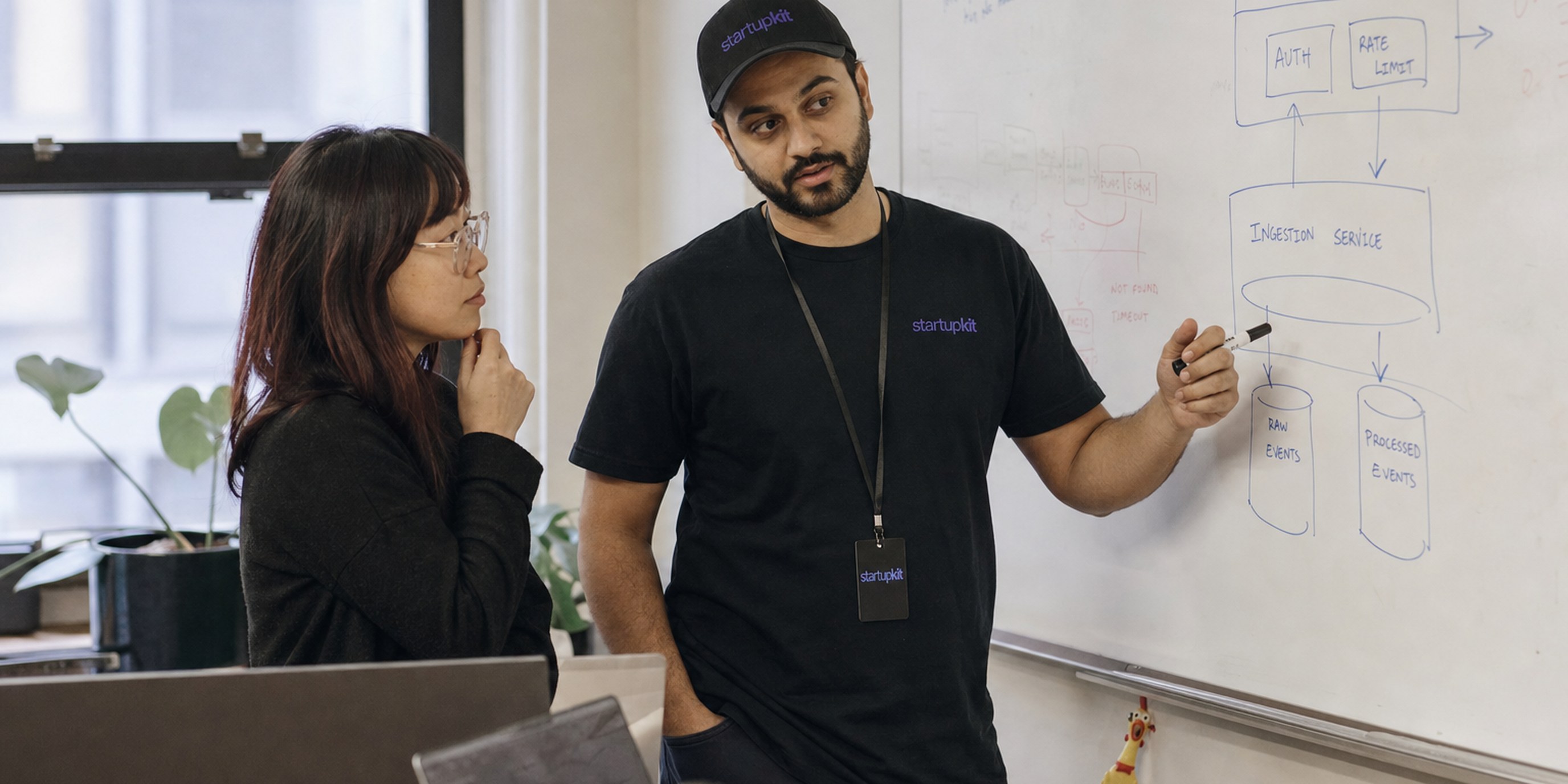 A startup founder and their first founding engineer reviewing a system design sketch on a whiteboard in a small office, laptops open beside them
