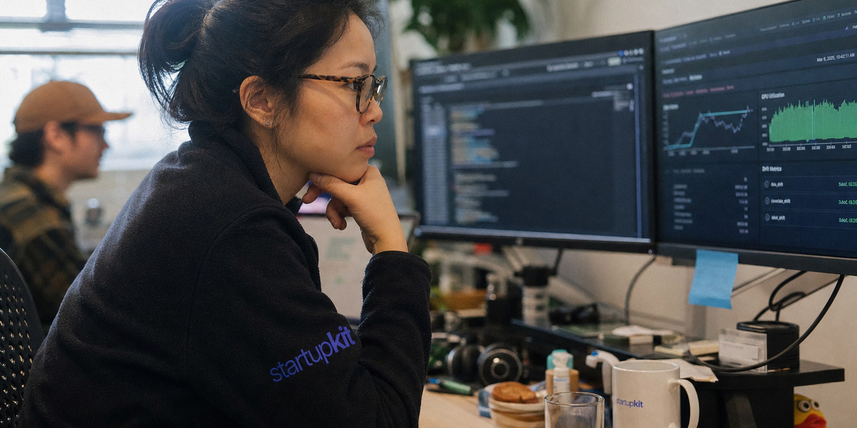 Machine learning engineer reviewing a model training run, drift metrics, and GPU utilization on dual monitors in a startup office