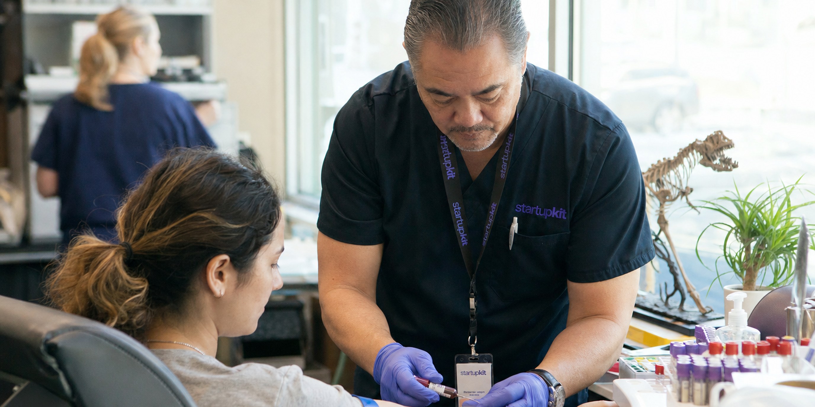 Male medical assistant in his fifties drawing blood from a seated patient's arm in a sunlit outpatient clinic