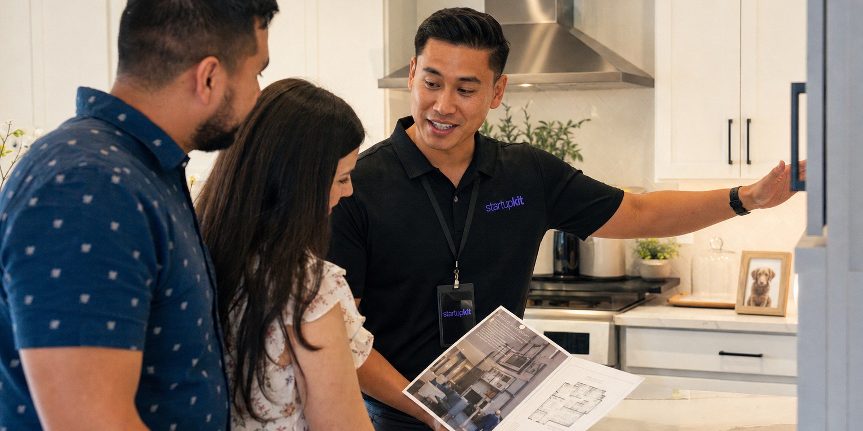 A new home sales specialist walking a young couple through a model-home kitchen, gesturing at the cabinetry as they review a floor-plan brochure