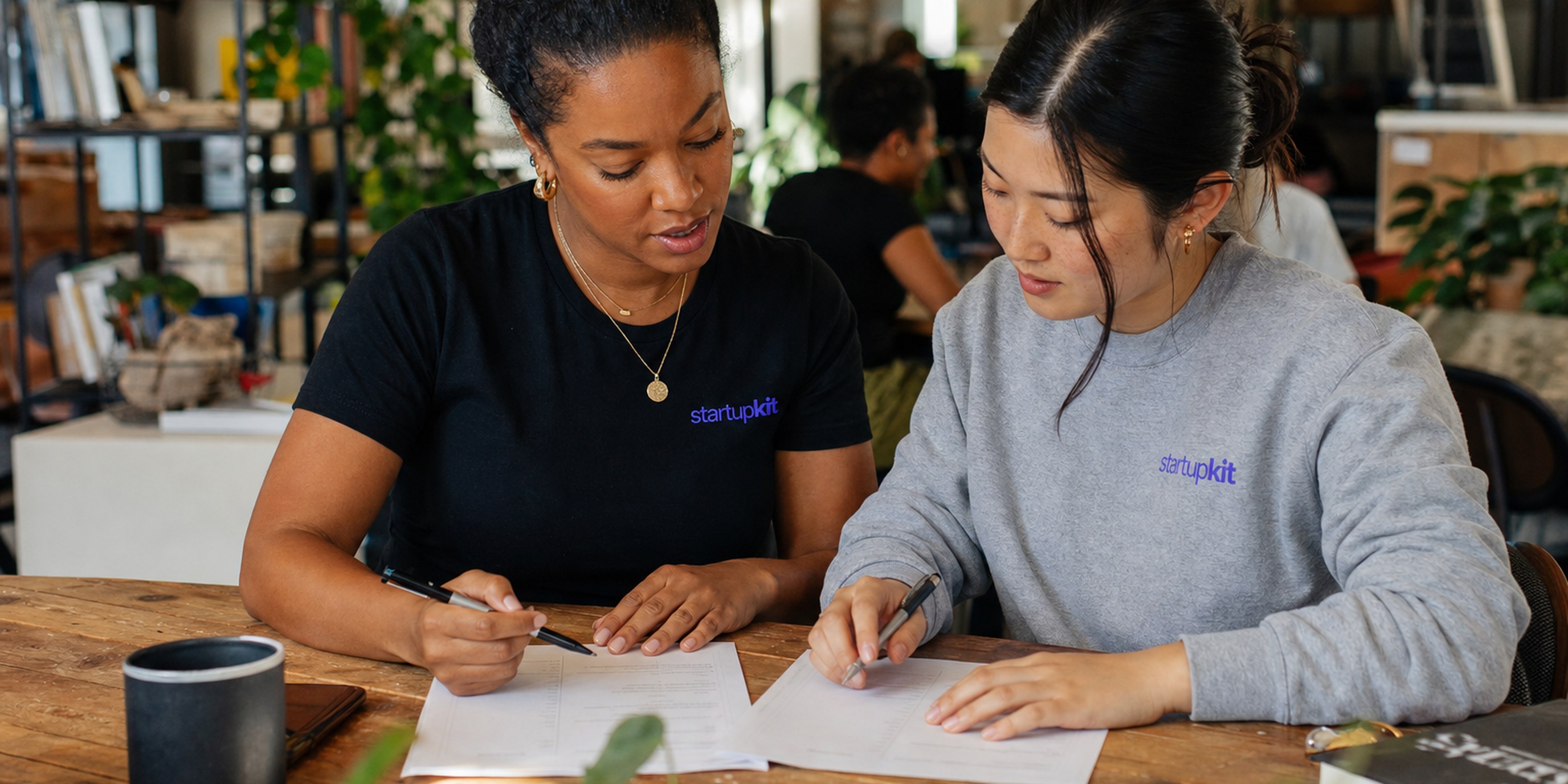 Two startup hiring managers comparing identical anchored interview rubrics side by side at a sunlit co-working table, each with their own independent ratings before the debrief