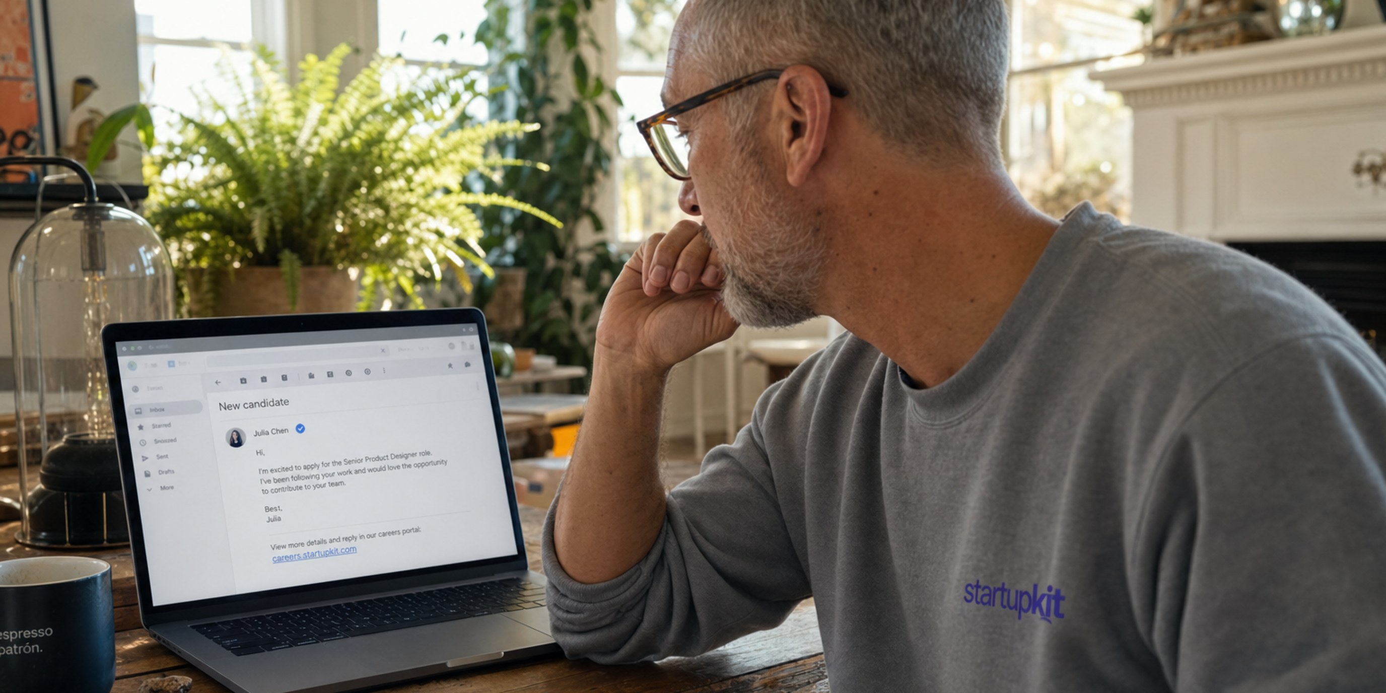 A recruiter in his late fifties at a sunlit home-office desk leaning toward a laptop that shows a candidate email with a verified-sender badge and a link to a branded company careers portal
